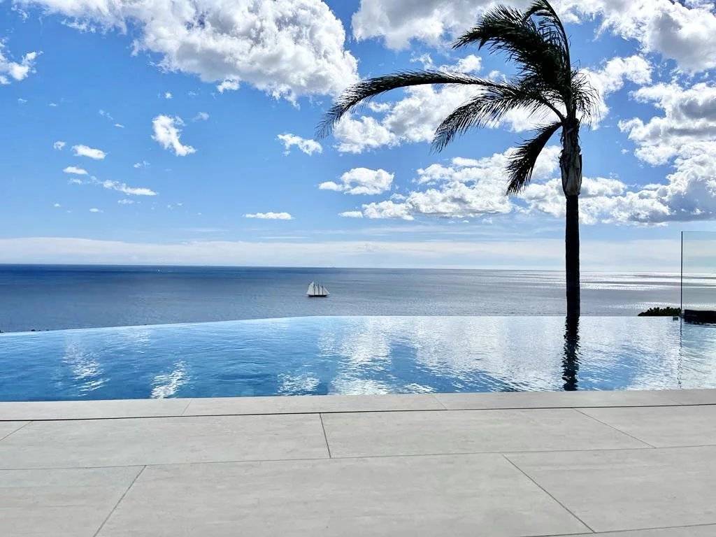 Piscine à débordement avec vue imprenable sur la mer Piscine à débordement, vue mer, palmier et voilier sous ciel bleu nuageux.